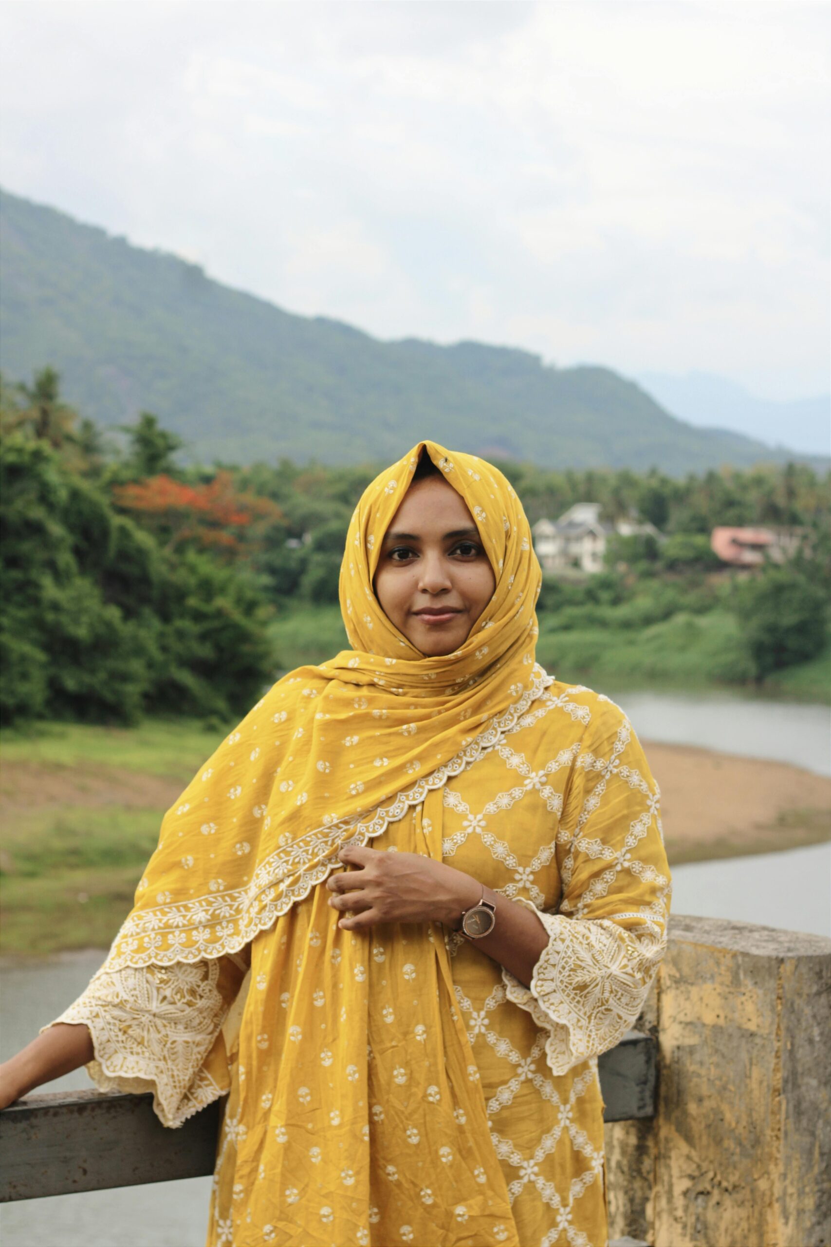 A serene portrait of a woman in a yellow dress and headscarf, standing by a river in Edavanna, India.